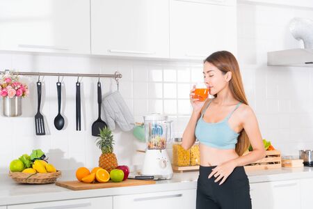 Asian woman wears a blue exercise suit and black pants is standing and drinking vegetable and fruit juice using blender on table in white kitchen.の写真素材