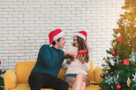 European couples, caucasian man wearing blue sweater and woman wearing white sweater, wearing christmas hat giving a Christmas present to his girlfriend in christmas season at sofa.の写真素材