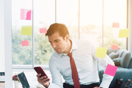Businessman wearing a white shirt with a red necktie sitting and watching mobile phone at office on background windows mirror with orange sunlight. Foreground glass meeting boardの写真素材