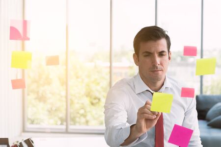 Businessman wearing a white shirt with a red necktie writing on post it at office on background windows mirror with orange sunlight. Foreground glass meeting board, Copy space left.の写真素材