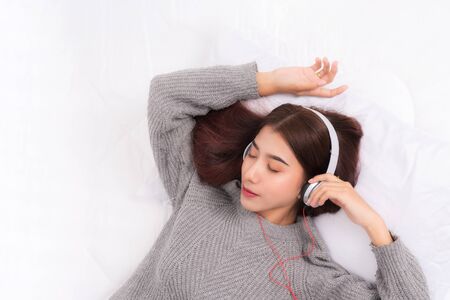 Young beautiful woman wearing a gray sweater is lying and listening to music wearing white music headphones on the bed. She is relaxing and down with eyes closed at happiness.の写真素材