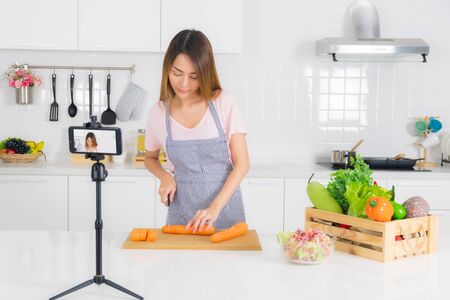 Asian beautiful woman, long hair, wearing a pink t-shirt and striped apron standing, slicing carrots on the table at modern white kitchen, and recording her cooking video using a mobile phone.の写真素材