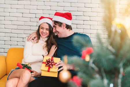 European couples, caucasian man wearing blue sweater and woman wearing white sweater, wearing christmas hat giving a Christmas present to his girlfriend in christmas season at sofa.の写真素材