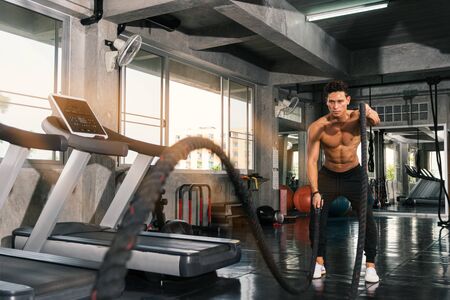 Athletic young man muscular powerful aggressive man training with rope in functional training fitness gym. in day time with sunlight at window.の写真素材
