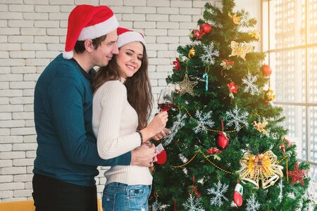 Celebration of the Christmas of couples. Young man are embracing their girlfriends, holding a glass of wine and enjoying standing beside the Christmas tree in the room.の写真素材