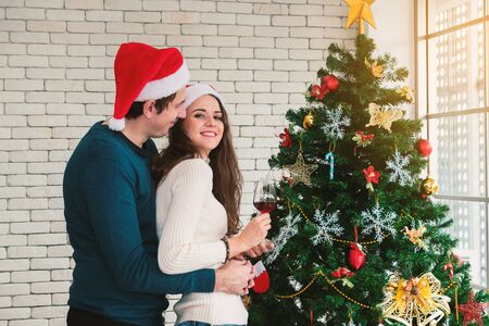 Celebration of the Christmas of couples. Young man are embracing their girlfriends, holding a glass of wine and enjoying standing beside the Christmas tree in the room.の写真素材