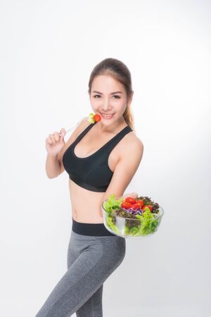 Asian woman portrait with perfect skin smiling face happy wearing black-gray exercise clothing holding a bowl of vegetable salad and fork on white background. Concept healthy.の写真素材