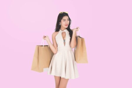 Happy beautiful asian woman brown long hair wearing a brown dress carrying shopping bags on isolated pink background, summer sale concept.の写真素材