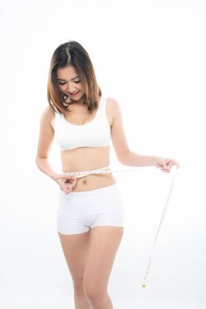 Young beautiful brunette woman wearing white underwear showing the measurements using measure tape at studio shoot isolated on white backgroundの写真素材