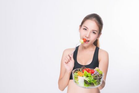 Asian woman portrait with perfect skin smiling face happy wearing black-gray exercise clothing holding a bowl of vegetable salad and fork on white background. Concept healthy.の写真素材