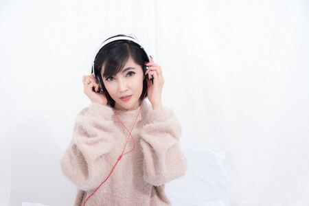 Beautiful woman long haired blond wearing sweater relaxing listening to music with headphones in the living room on white curtain background. She is relaxing and happiness.の写真素材