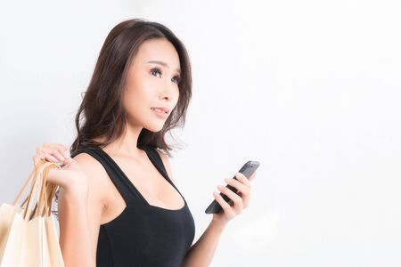 Happy beautiful asian woman black long-haired woman wearing a black shirt, jeans using a mobile phone and carrying shopping bags  on isolated gray background, Shopping summer sale concept.の写真素材