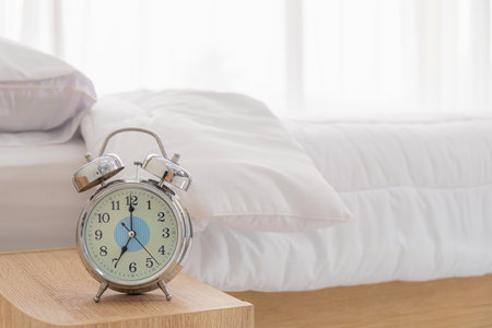 Close Up view. Alarm clock is located on the table at the head on white bed decoration in bedroom.