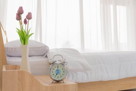 Close Up view. Alarm clock and Tulips flower vase is located on the table at the head on white bed decoration in bedroom.の写真素材