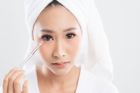 Beautiful young woman wearing bathrobe with towel with towel on head is using a makeup brush makeup her after bathing, at studio shot isolated on white background.の写真素材