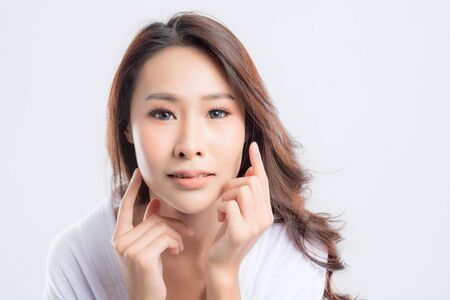 Beautiful young woman wearing white bathrobes is touching her face and smiling isolated on white background. Woman after bath with clean perfect skin.の写真素材