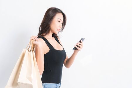 Happy beautiful asian woman black long-haired woman wearing a black shirt, jeans using a mobile phone and carrying shopping bags  on isolated gray background, Shopping summer sale concept.の写真素材