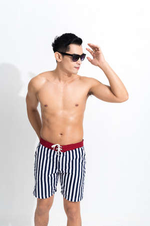 Young muscular guy in striped beach shorts sunbathing wear sunglasses at studio shot isolated on white background. Fashion summer concept.の写真素材