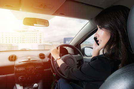 Beautiful asian woman wear black suit work clothes talking on a mobile phone at car in evening time on sunlight background.の写真素材