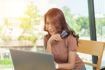 Beautiful woman wearing a brown shirt sitting and smiling face happy sitting by a large window, She is shopping online with laptop and holding credit card.の写真素材