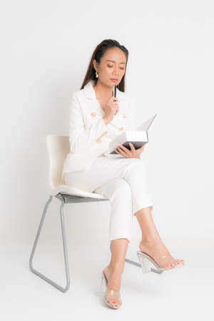 Fashion style catalog clothing for business woman black long hair natural make up wear white suit sitting on white chair, holding book and pens at studio shoot on white background.の写真素材