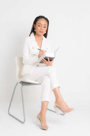 Fashion style catalog clothing for business woman black long hair natural make up wear white suit sitting on white chair, holding book and pens at studio shoot on white background.の写真素材