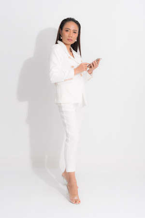 Fashion style catalog clothing for business woman black long hair natural make up wear white suit costume perfect body shape suit, using mobile phone at studio shoot on white background and shadow.の写真素材