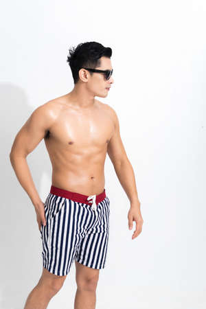 Young muscular guy in striped beach shorts sunbathing wear sunglasses at studio shot isolated on white background. Fashion summer concept.の写真素材