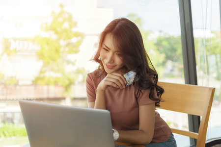 Beautiful woman wearing a brown shirt sitting and smiling face happy sitting by a large window, She is shopping online with laptop and holding credit card.の写真素材