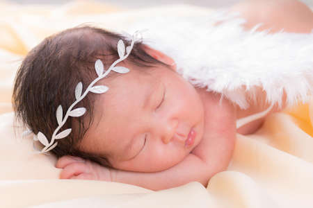 Portrait of a one month old sleeping, newborn baby girl. She is wearing a white crown headband, angel wings and sleeping on a cream blanket. Concept portrait studio fashion newborn.の写真素材