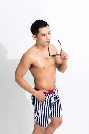 Young muscular guy in striped beach shorts sunbathing, holding sunglasses at studio shot isolated on white background. Fashion summer concept.の写真素材