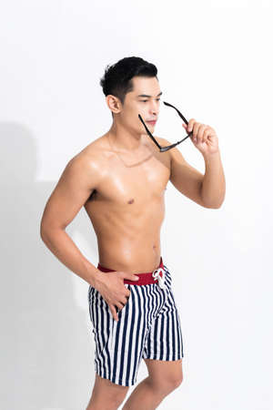 Young muscular guy in striped beach shorts sunbathing, holding sunglasses at studio shot isolated on white background. Fashion summer concept.の写真素材