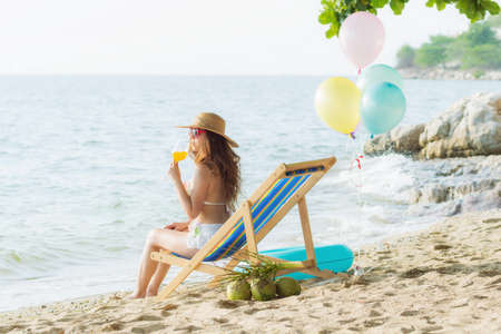 Happy woman in swimsuit holding cocktail, sitting in lounge chair on beach , banner design with empty space. Woman having relaxing summer vacation at beach resortの写真素材