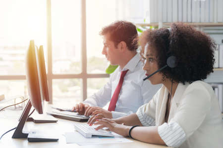 Woman call center African American wearing white work clothes with headset working in call center at computer.の写真素材
