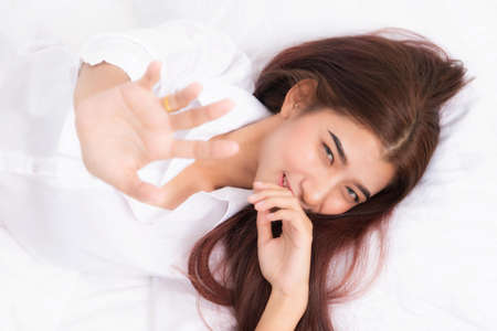 Cheerful young woman lying on bed playing with blanket and looking at camera with white shirt at the white bed in the morning.の写真素材