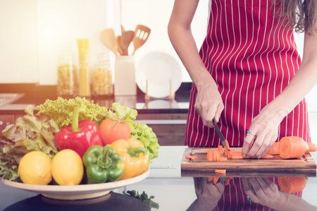 Close-up woman in a red apron precisely chops fresh carrots on a wooden board, with a vibrant bowl...