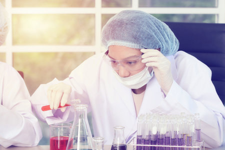 Focused female scientist in a white lab coat, wearing a hairnet, mask, and safety glasses,...