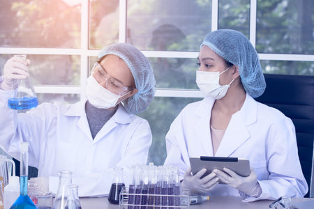 Two female scientists in lab coats and protective gear are performing an experiment, holding a flask...
