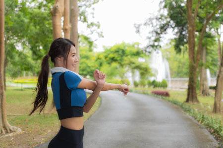 Side view of a fit Asian woman doing arm stretches in a beautiful park setting, preparing for her outdoor fitness routine.の写真素材