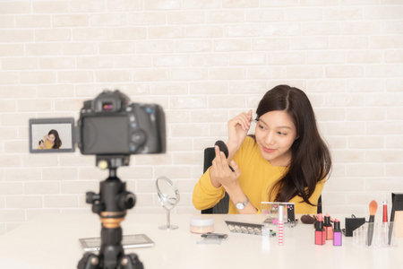 young Asian woman records a beauty vlog with camera, demonstrating how to curl her eyelashes while looking into a mirror.の写真素材