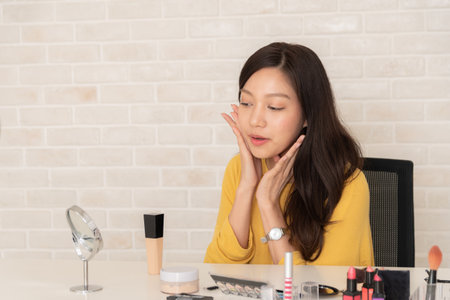 Young Asian woman happily applies makeup or skincare to her face, looking in a mirror with various beauty products on a white desk.の写真素材