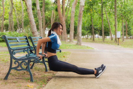 Smiling Asian woman performs triceps dips using a park bench, demonstrating outdoor exercise and workout routine.の写真素材