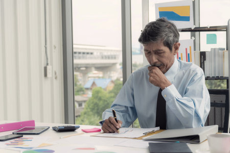 Experienced elderly Asian businessman in a blue shirt and tie intently analyzing financial charts...
