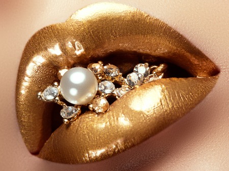 Cosmetics and make-up. Closeup of gold artistic lips. Glitter shiny lip makeup. Shoot of a beautiful girl with golden lipstick and gloss. Sexy and stylish lips. Pearls and diamonds in her mouthの写真素材
