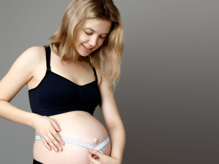 Pregnant woman measuring her belly while standing against a white background. Smiling pregnant woman measuring her large belly, on gray backgroundの写真素材