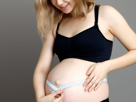 Pregnant woman measuring her belly while standing against a white background. Smiling pregnant woman...