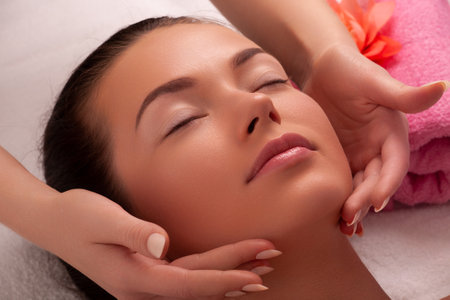 Beautiful caucasian woman enjoying facial massage with closed eyes in sunny spa salon. Relaxing treatment in medicineの写真素材
