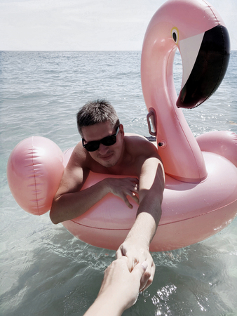 Young man swimming on inflatable pink flamingo in a sea at summer vacationの写真素材