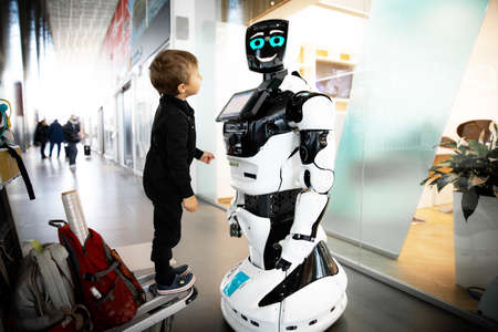 Small boy of European appearance in a black overalls stands on a trolley for suitcases and looks with interest at a robot that is standing and smiling in a public place.の写真素材