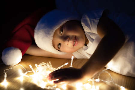 A little boy in a Santa hat and a white T-shirt lies on the floor and plays in the dark with Christmas garlands. The kid smiles sweetly and is waiting for the New Years holiday.の写真素材